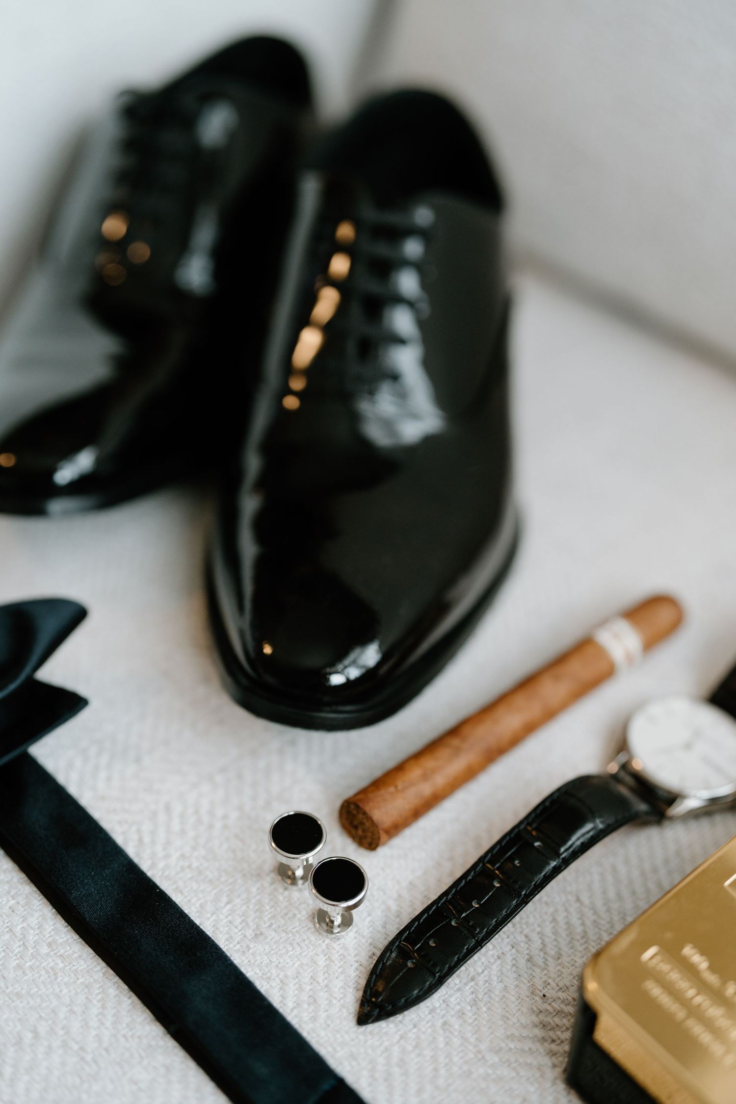 Black patent leather dress shoes with a brown cigar and gold accessories arranged on a white textured surface.