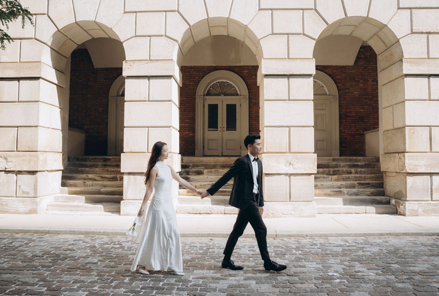 Two people in wedding attire walk hand in hand under elegant stone archways of a classical building.