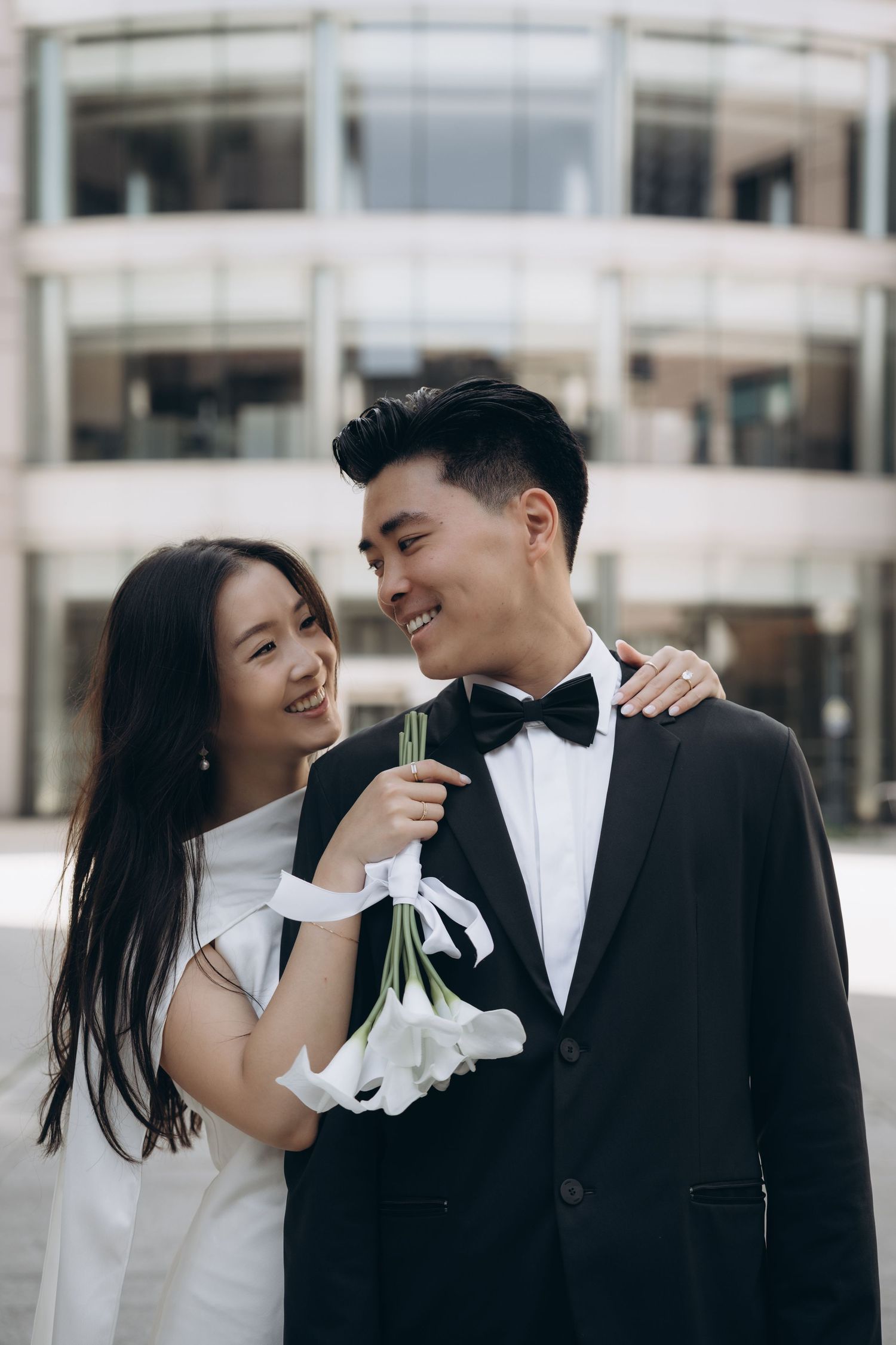 A couple in wedding attire share a joyful moment together outside a modern building.