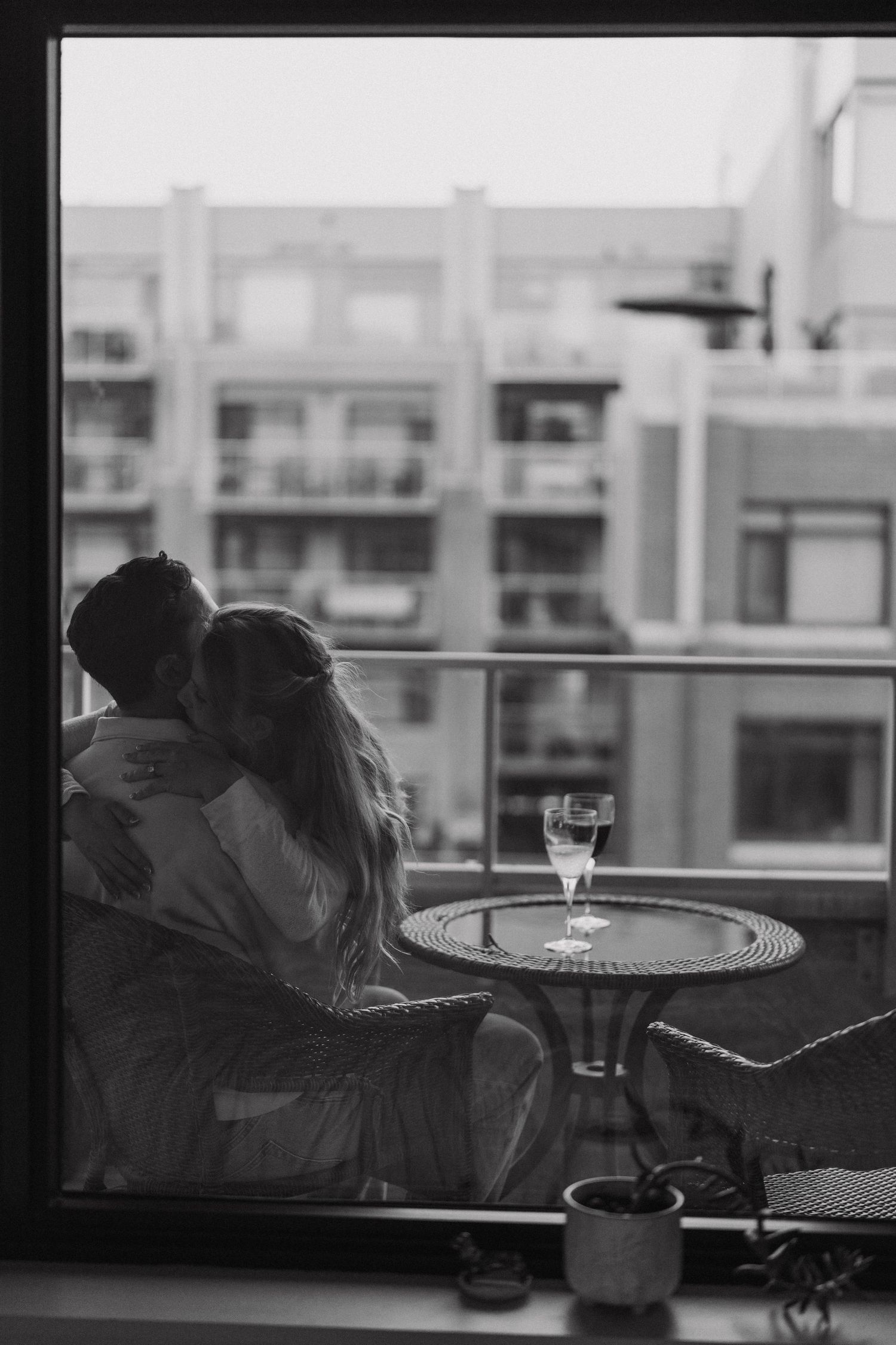 A romantic silhouette of a couple embracing by a window with a glass of wine on a table in black and white photography.