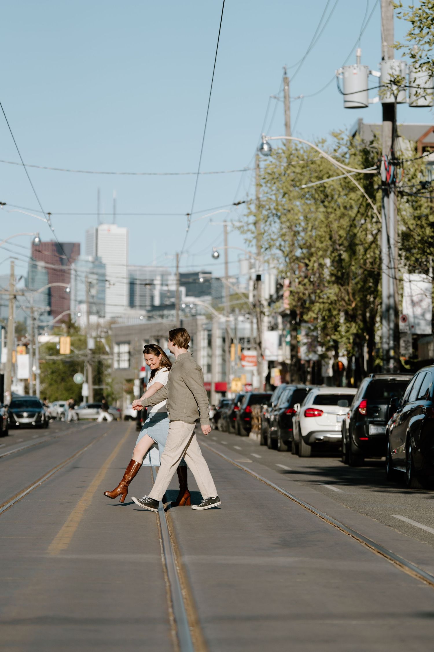 Couple crossing a street with light rail tracks in an urban setting with parked cars and skyscrapers in the background.