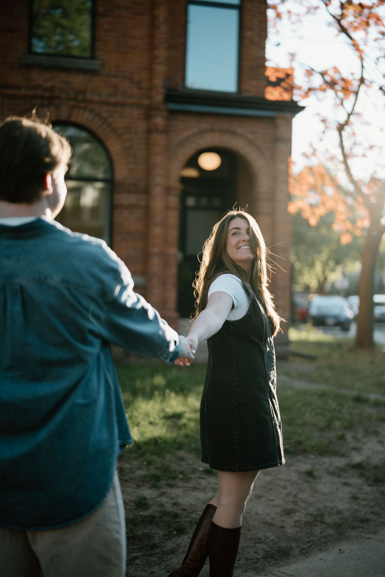 Couple holding hands in fall sunshine outside brick building as woman in dress and boots spins playfully in golden light.