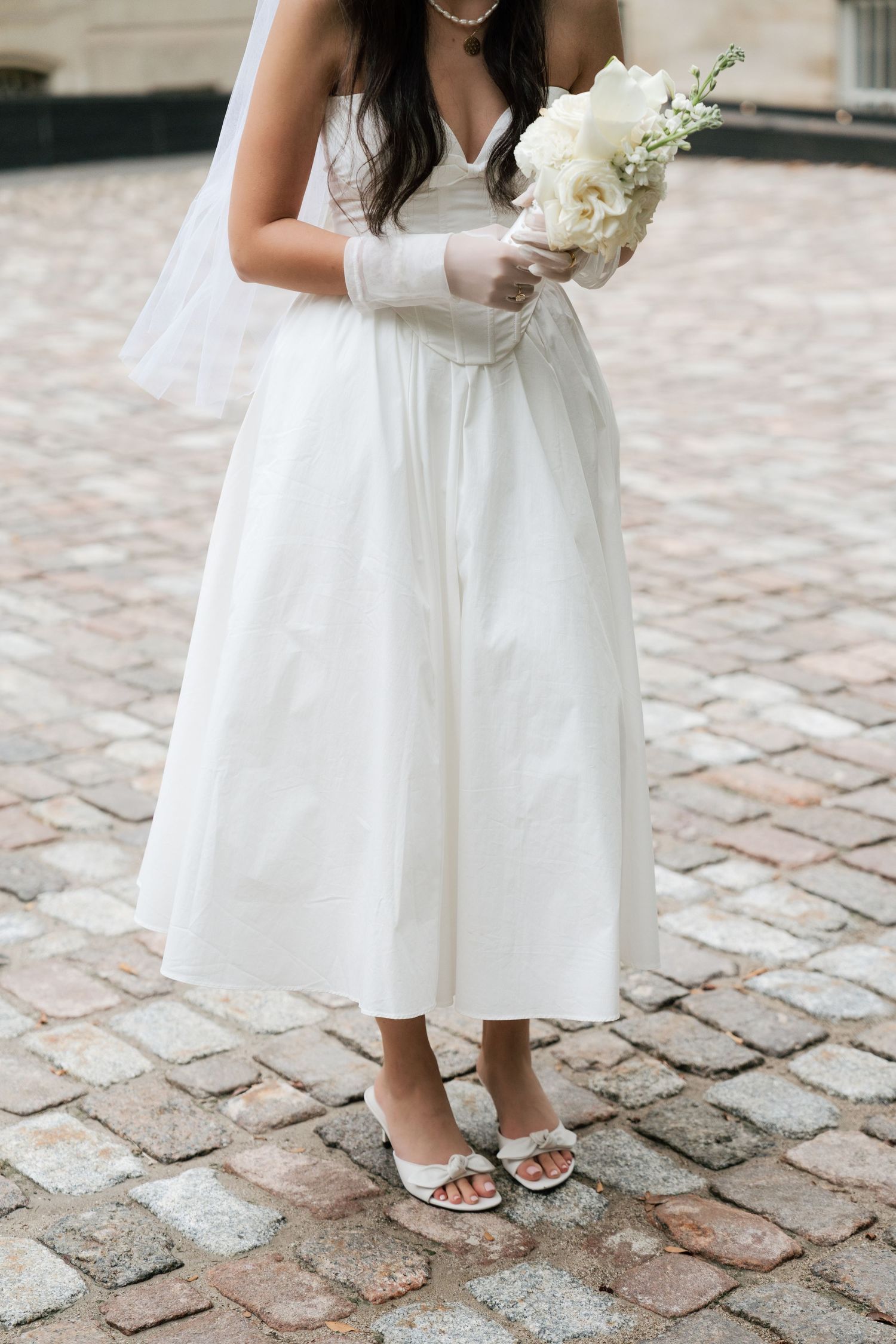 Close-up view of white dress and bouquet details on cobblestone pavement during a wedding ceremony.