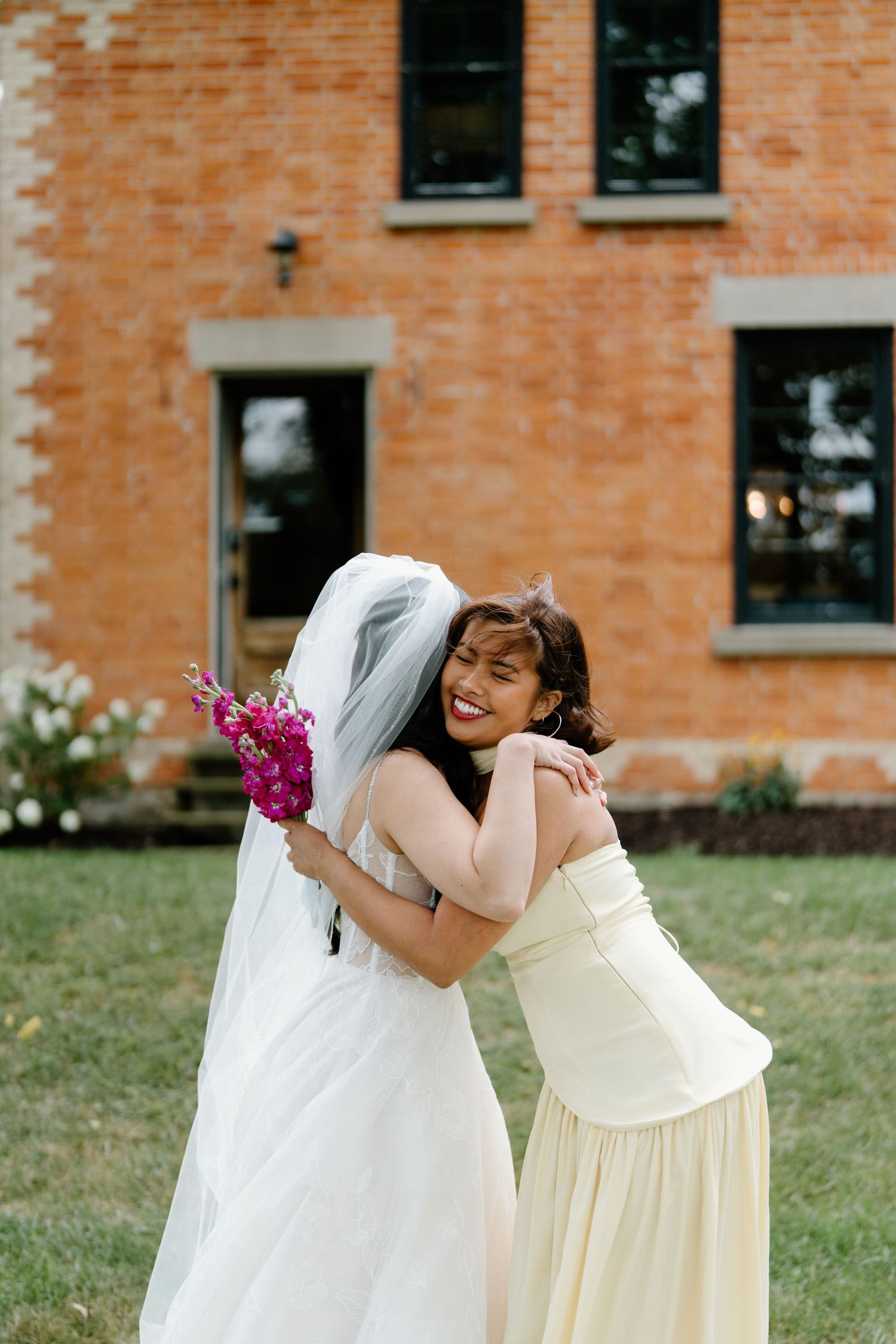 Two women in cream and white dresses share a joyful embrace outdoors on a grassy lawn with brick building behind them.