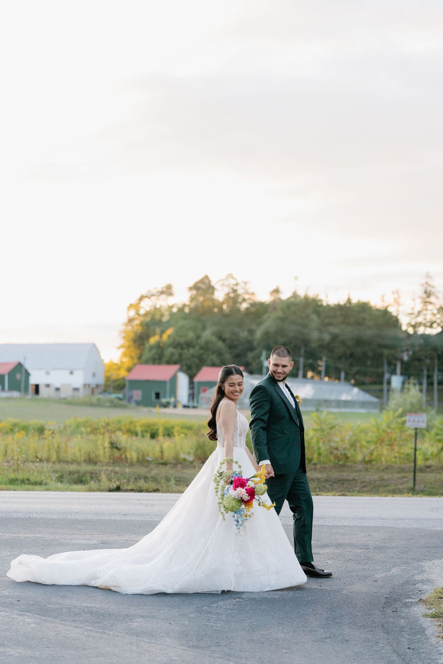 A couple in wedding attire creates romantic poses during their outdoor photoshoot near rural buildings at dusk.