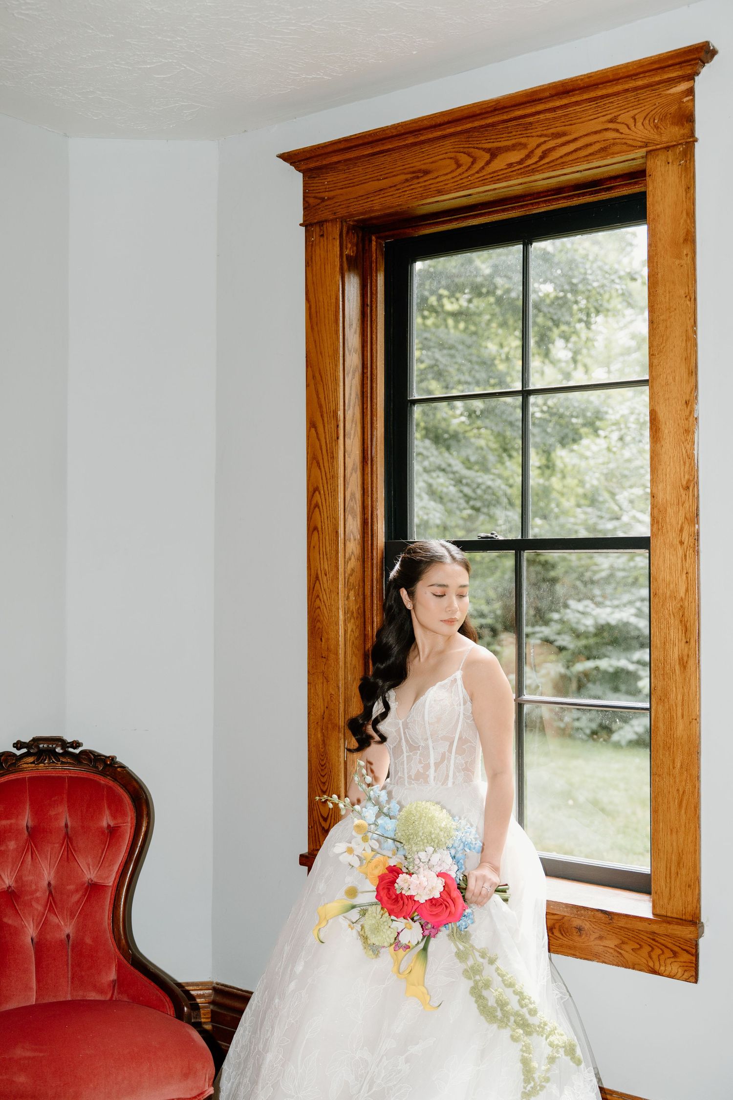 A bride in a white wedding dress sits elegantly by a window in a room with red velvet seating and wooden trim.