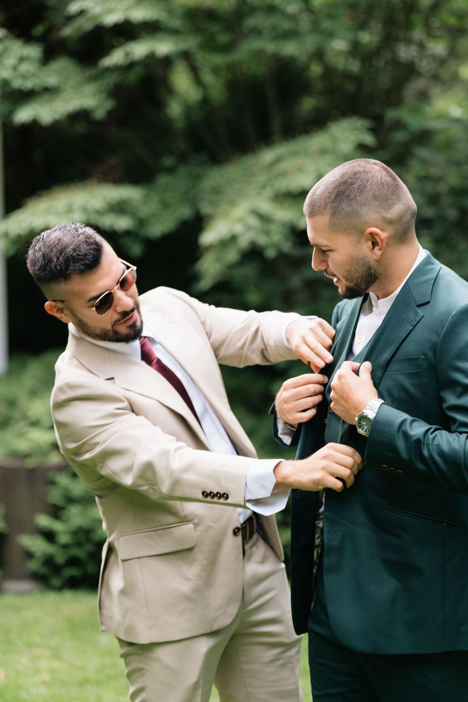 Two men in contrasting suits playfully adjust each other's boutonnieres in a garden setting.