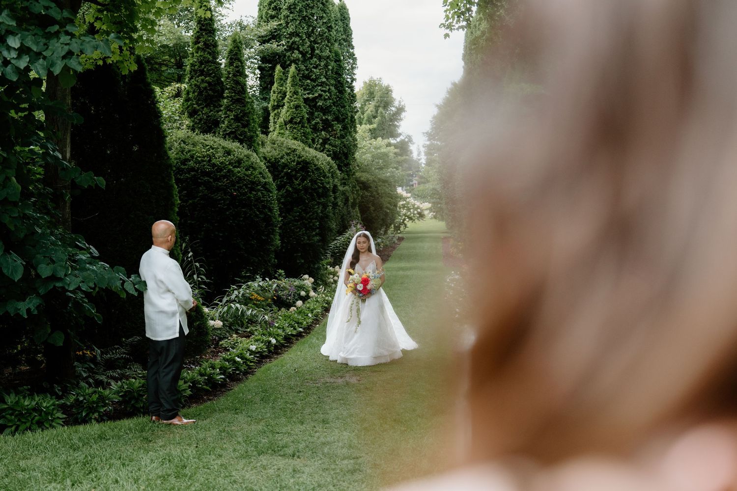 A wedding photographer captures a moment in a lush garden path lined with manicured hedges and greenery.
