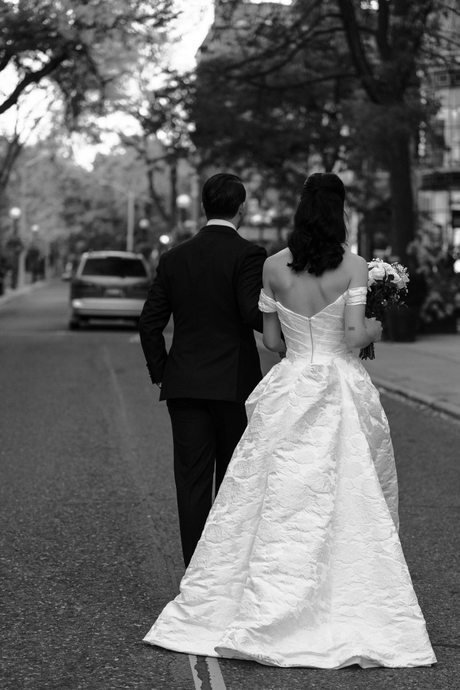 A couple walks down a tree-lined street after their wedding, with the bride wearing a flowing white off-shoulder gown.