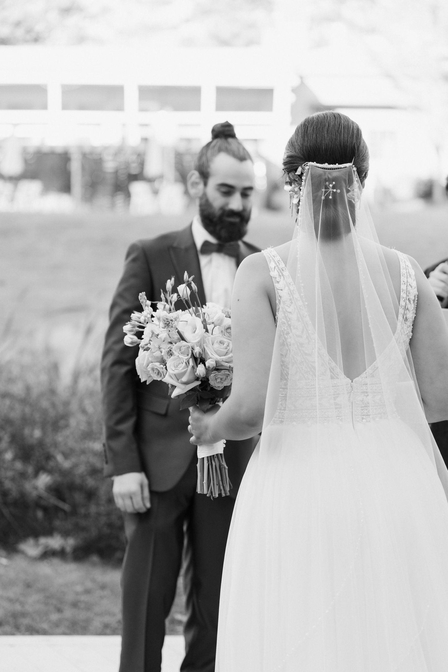 Black and white photo of a bride holding a bouquet while facing her groom in formal wedding attire.