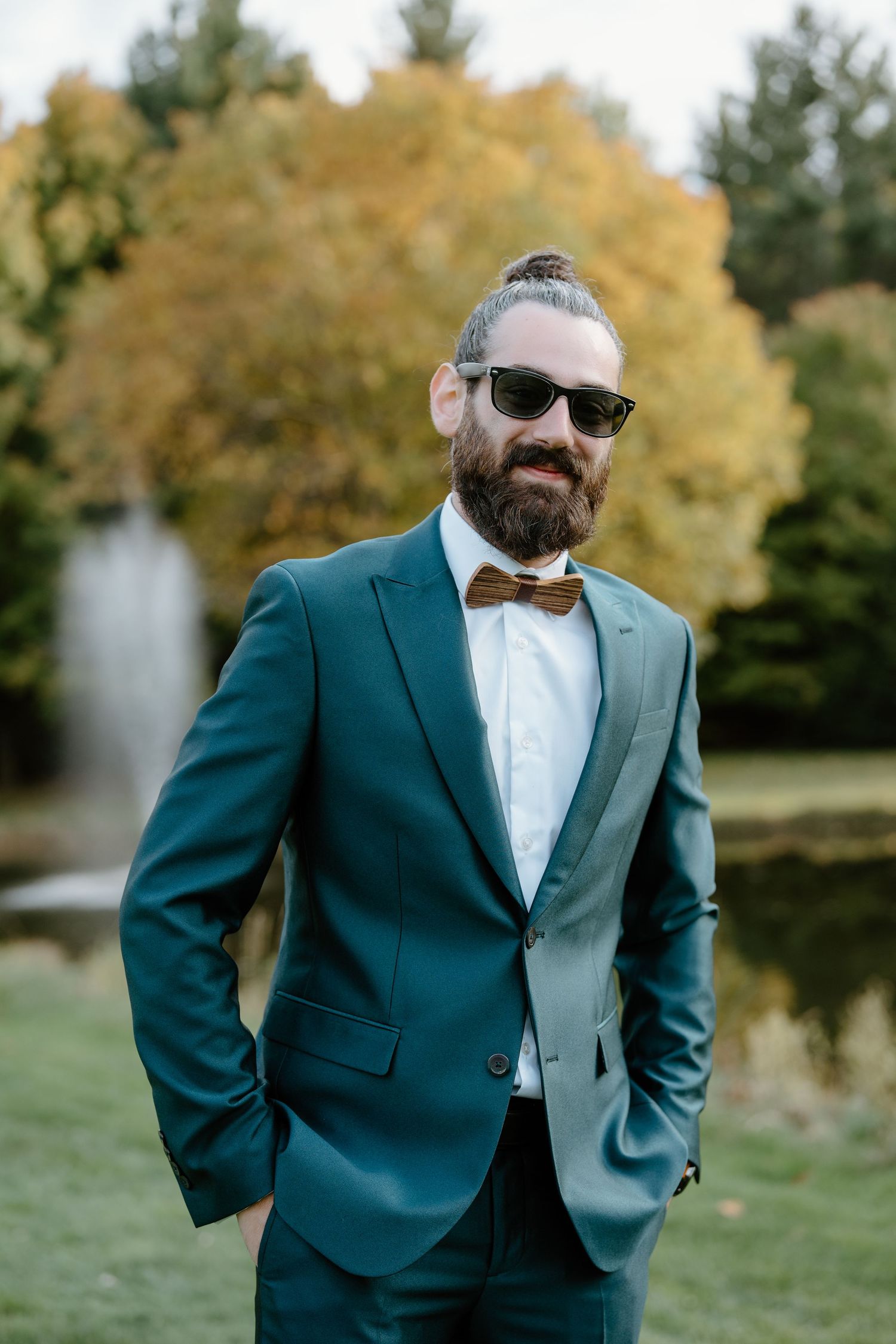 Person in teal suit and wooden bow tie poses outdoors with autumn foliage background.
