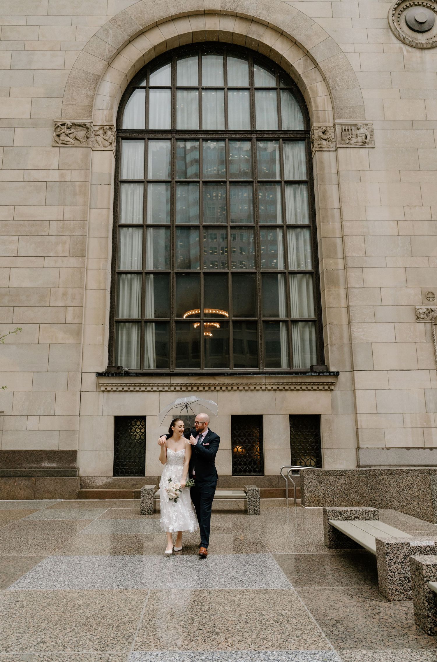 A couple stands outside a grand stone building with a large arched window and ornate architectural details.