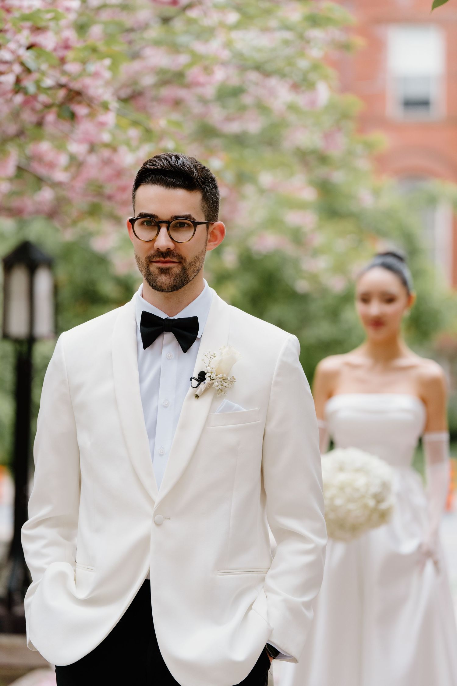 Elegant wedding scene with white tuxedo and bow tie against pink cherry blossoms and brick building background.