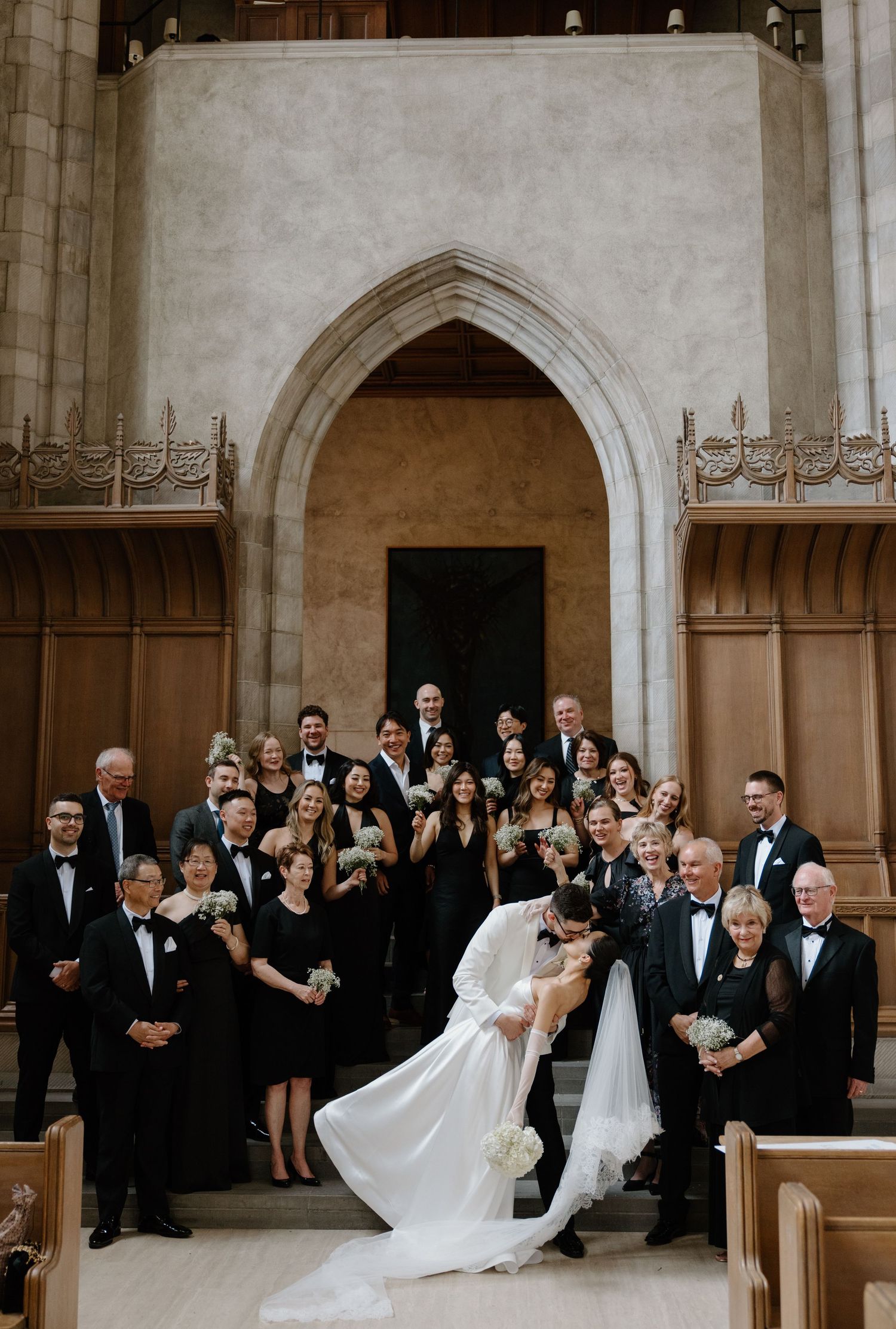 Wedding party poses for formal group photo under gothic church archway in black and white attire.
