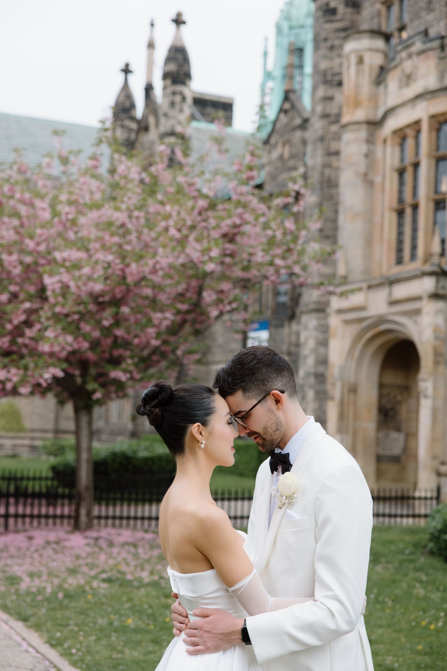 A romantic couple embraces in front of a Gothic stone building with blooming cherry blossom trees in spring.