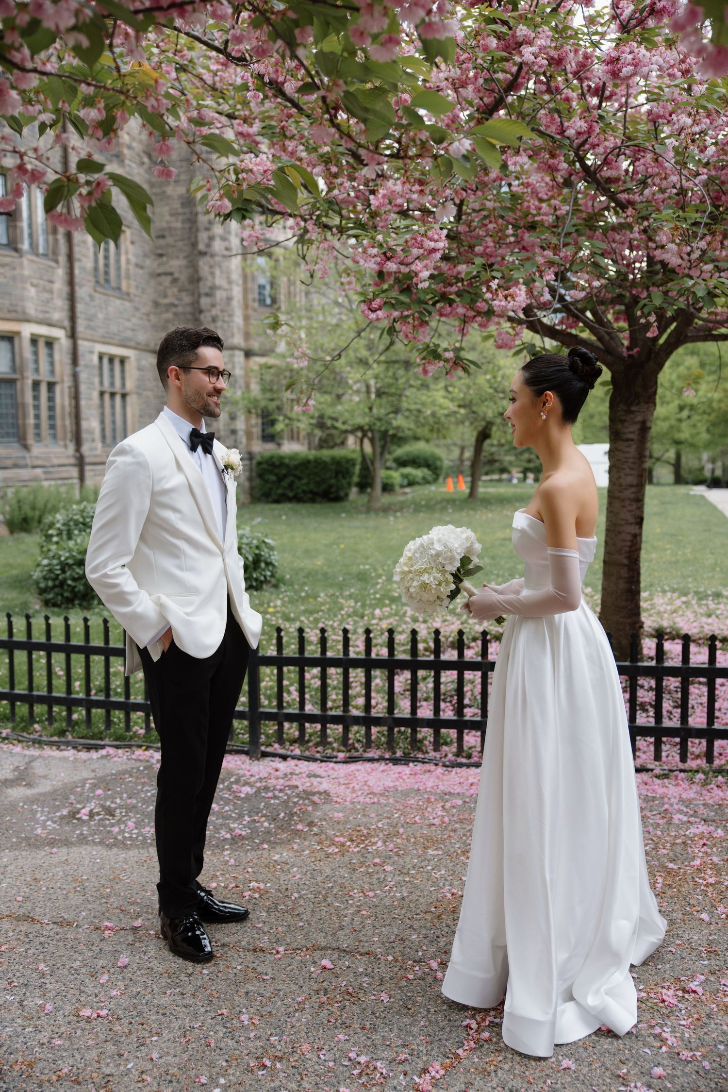 A bride and groom share a moment under blooming cherry blossoms near a stone building and iron fence.