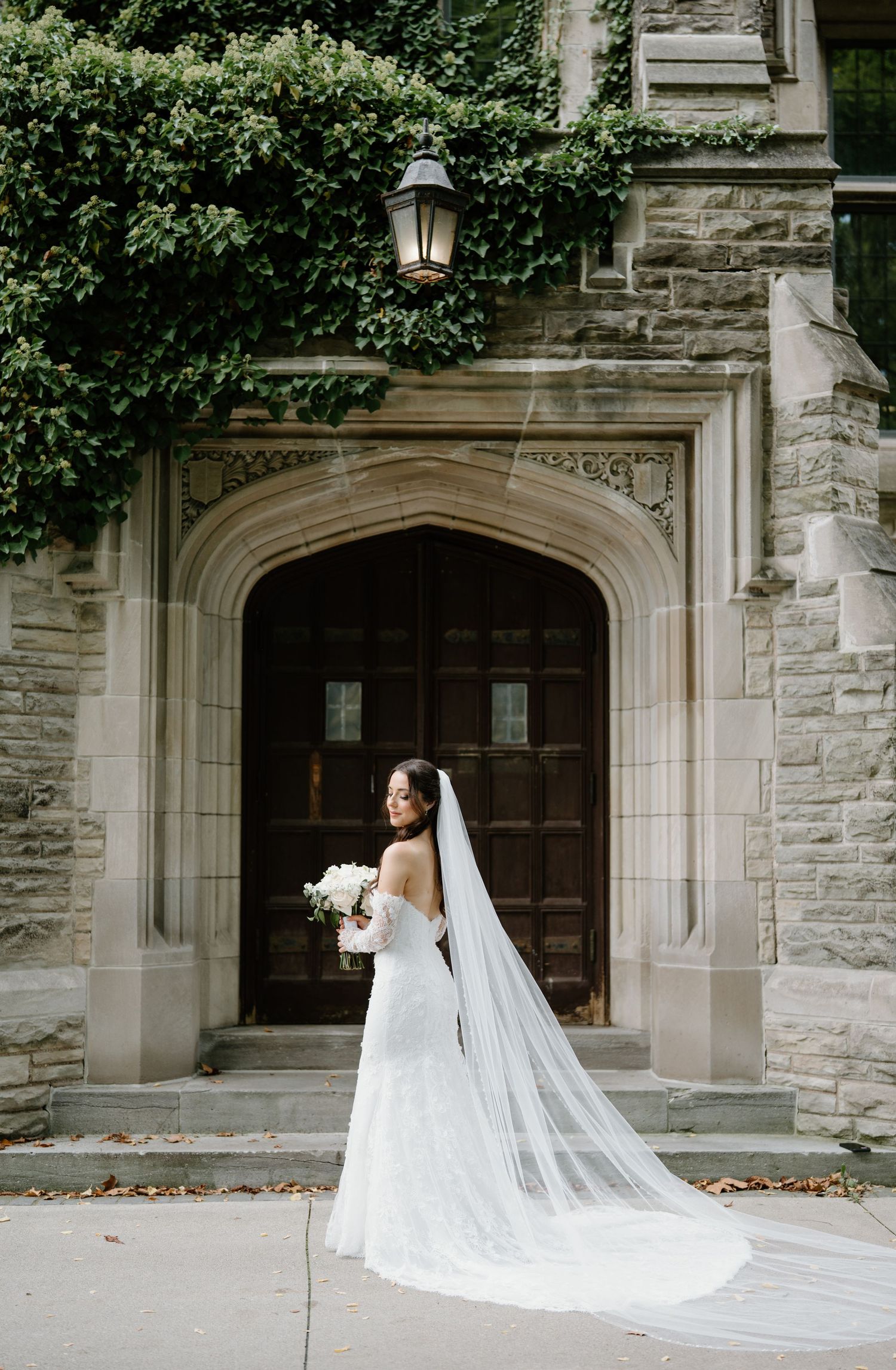 A flowing cathedral length veil trails behind a bride in a white gown against a stone church entrance.