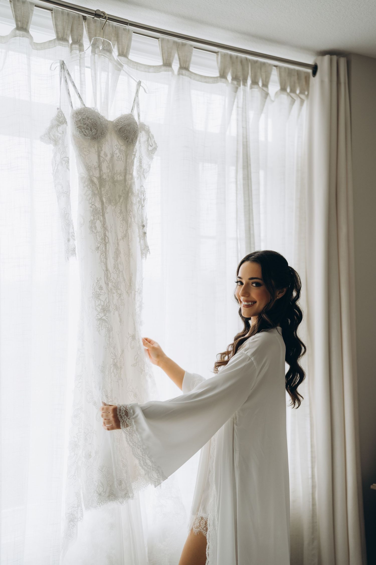 A person in a white robe admiring a wedding dress hanging by a window with natural light.
