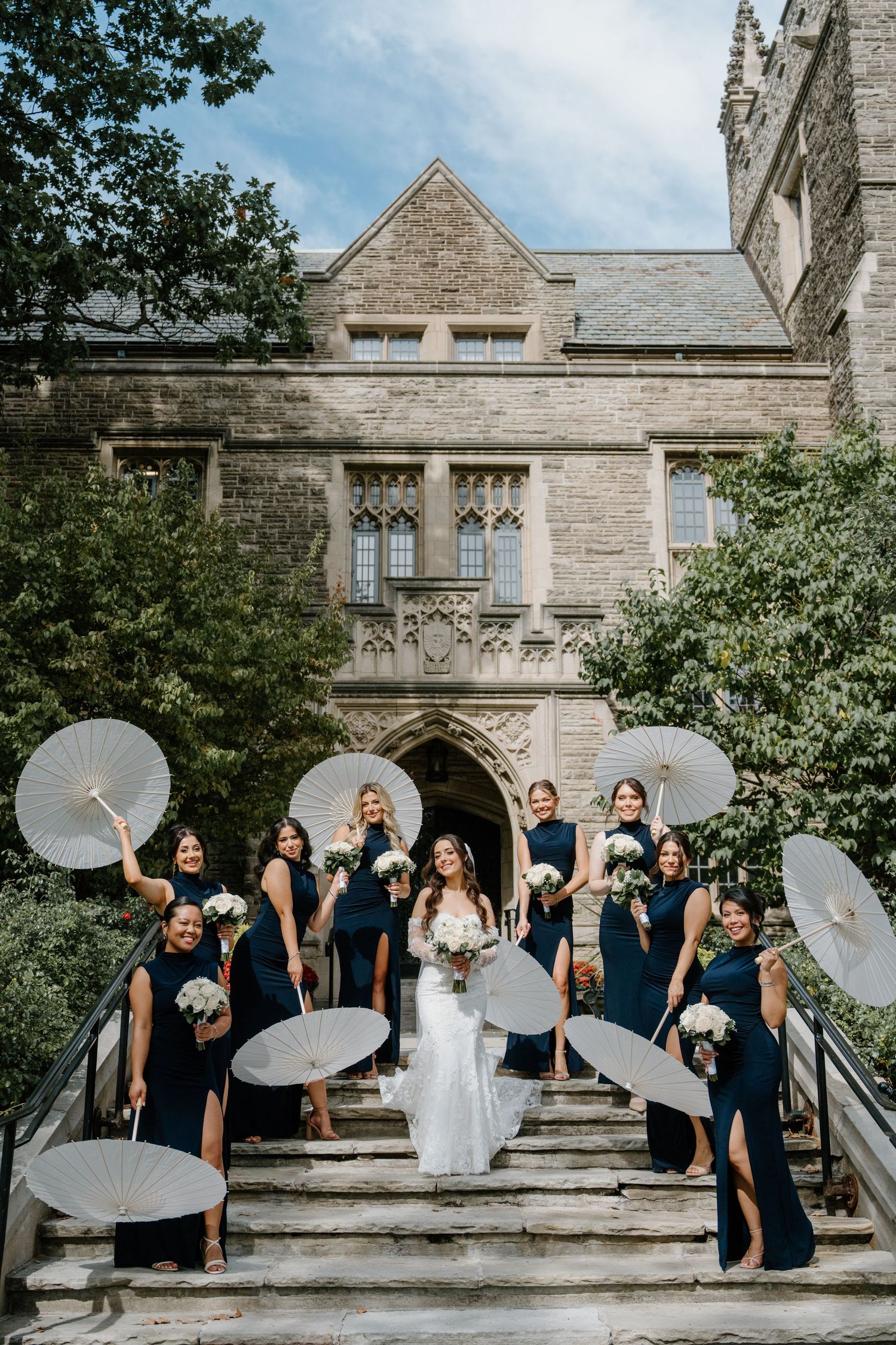 Wedding party with white parasols creates dynamic poses in front of historic stone church exterior.