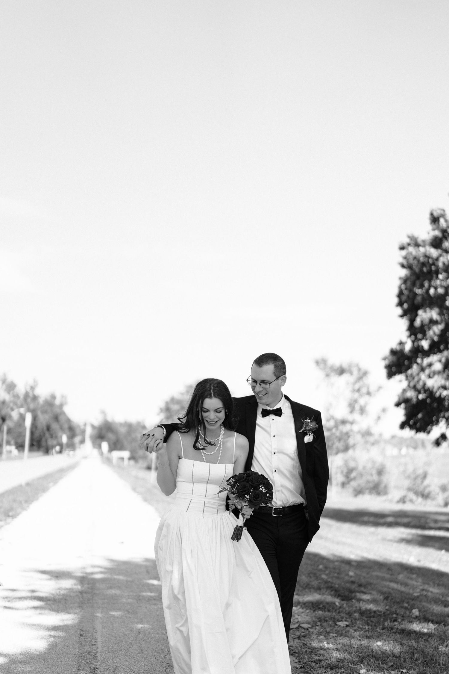 A joyful couple strolls hand in hand on a sunny path through a serene outdoor setting in formal attire.