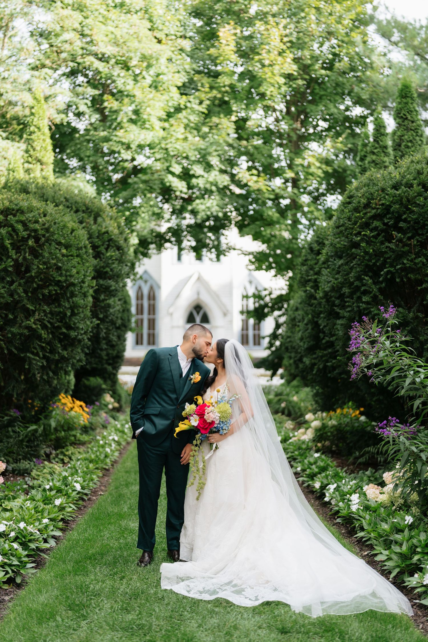 A couple shares an intimate moment in a lush garden path lined with greenery and flowers near a church.