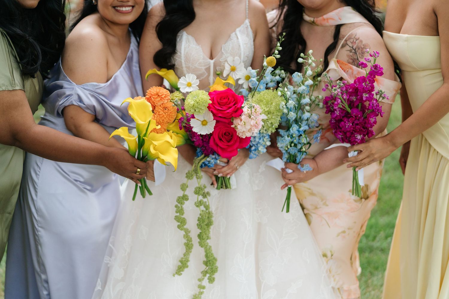 Wedding party holding vibrant multicolored bouquets featuring roses, lilies and cascading greenery against outdoor setting.