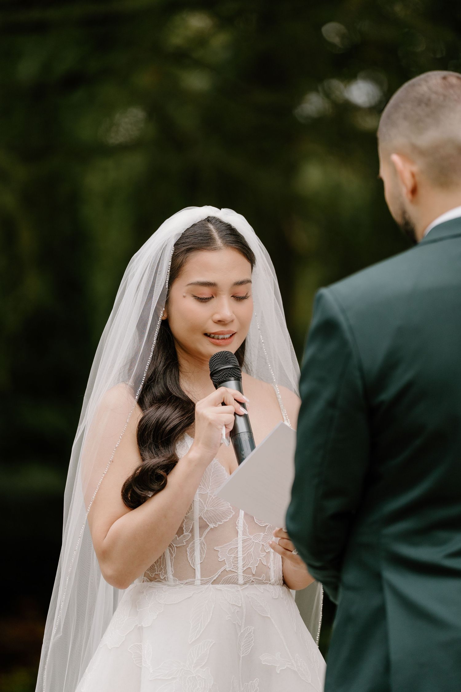 A bride in a white wedding dress and veil laughs joyfully during an outdoor ceremony.