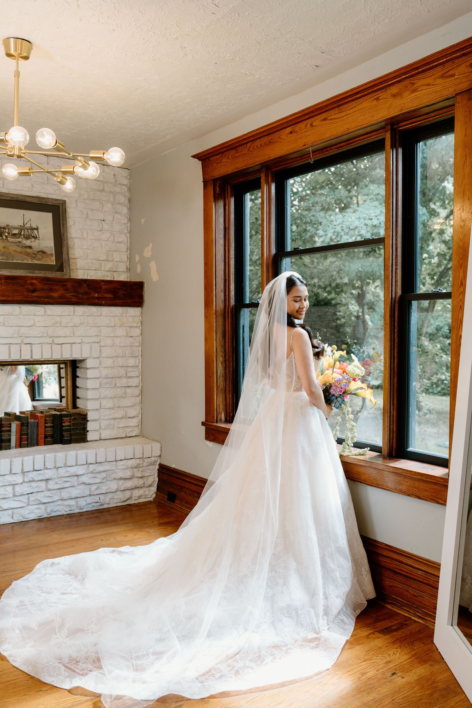 A flowing wedding gown with cathedral train displayed in a room with white brick walls and wooden windows.