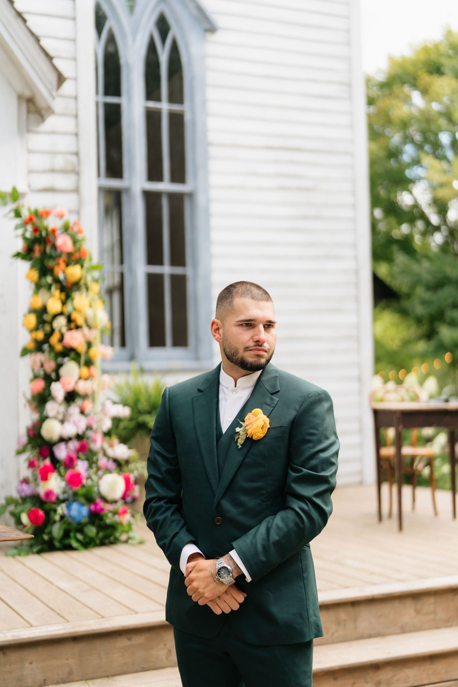 A person in a dark suit stands on a white church porch decorated with colorful floral arrangements.