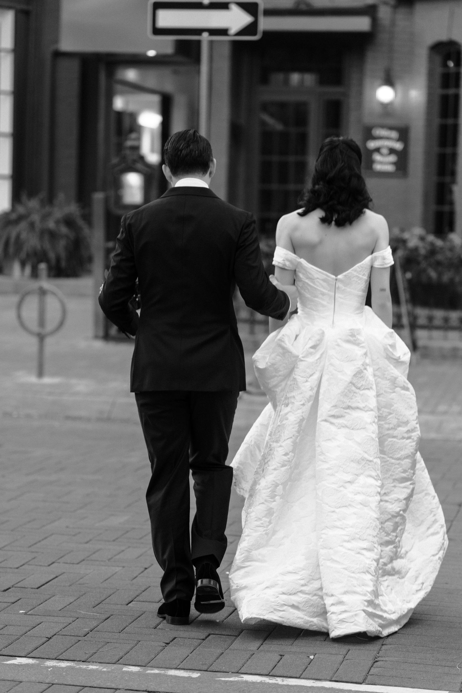 A bride in a white off-shoulder gown and groom in black suit walk together on a brick sidewalk in black and white.
