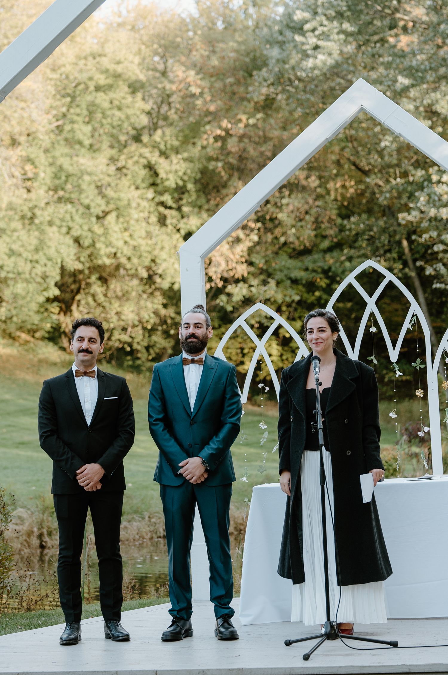 Three officiants stand at a modern white wedding arch during an outdoor ceremony.