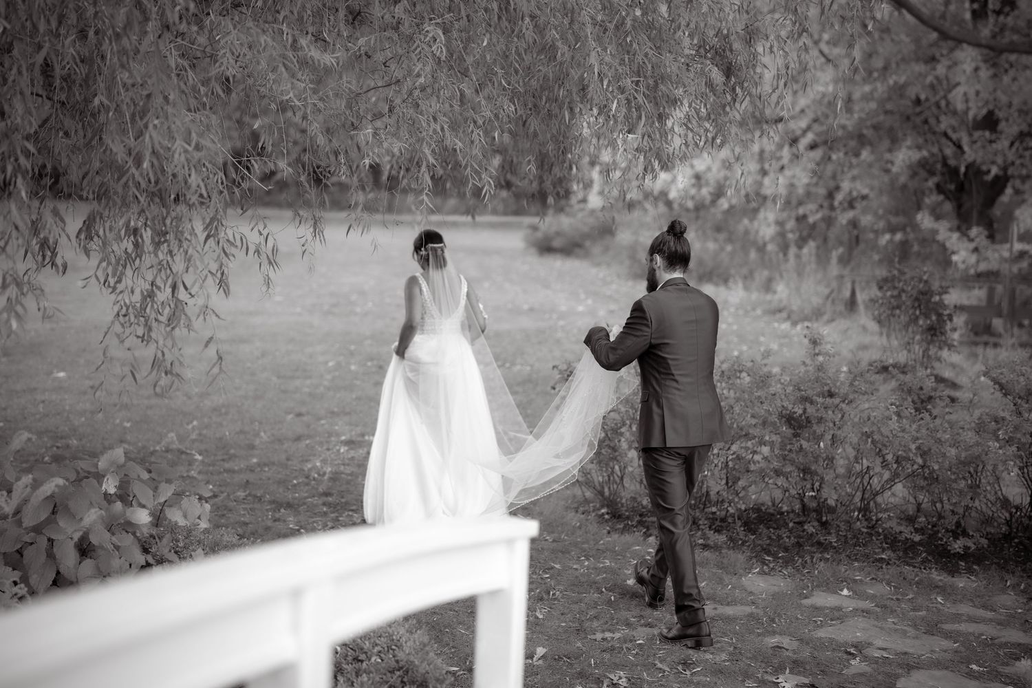 Black and white shot of bride in flowing dress walking on wooden bridge with photographer.