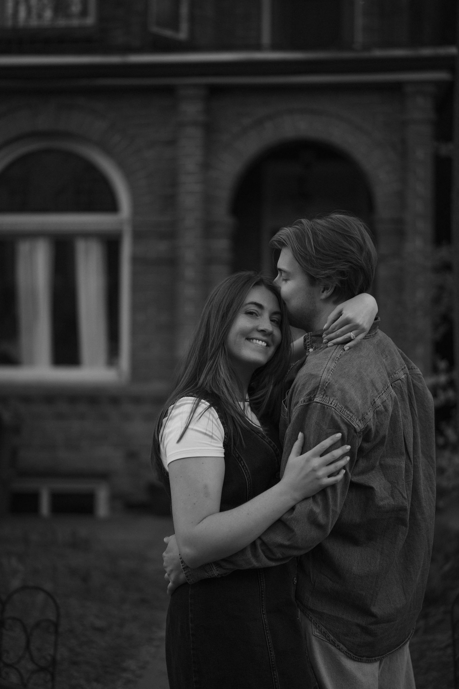 A couple shares an intimate embrace outside a brick building with arched windows in a black and white photograph.