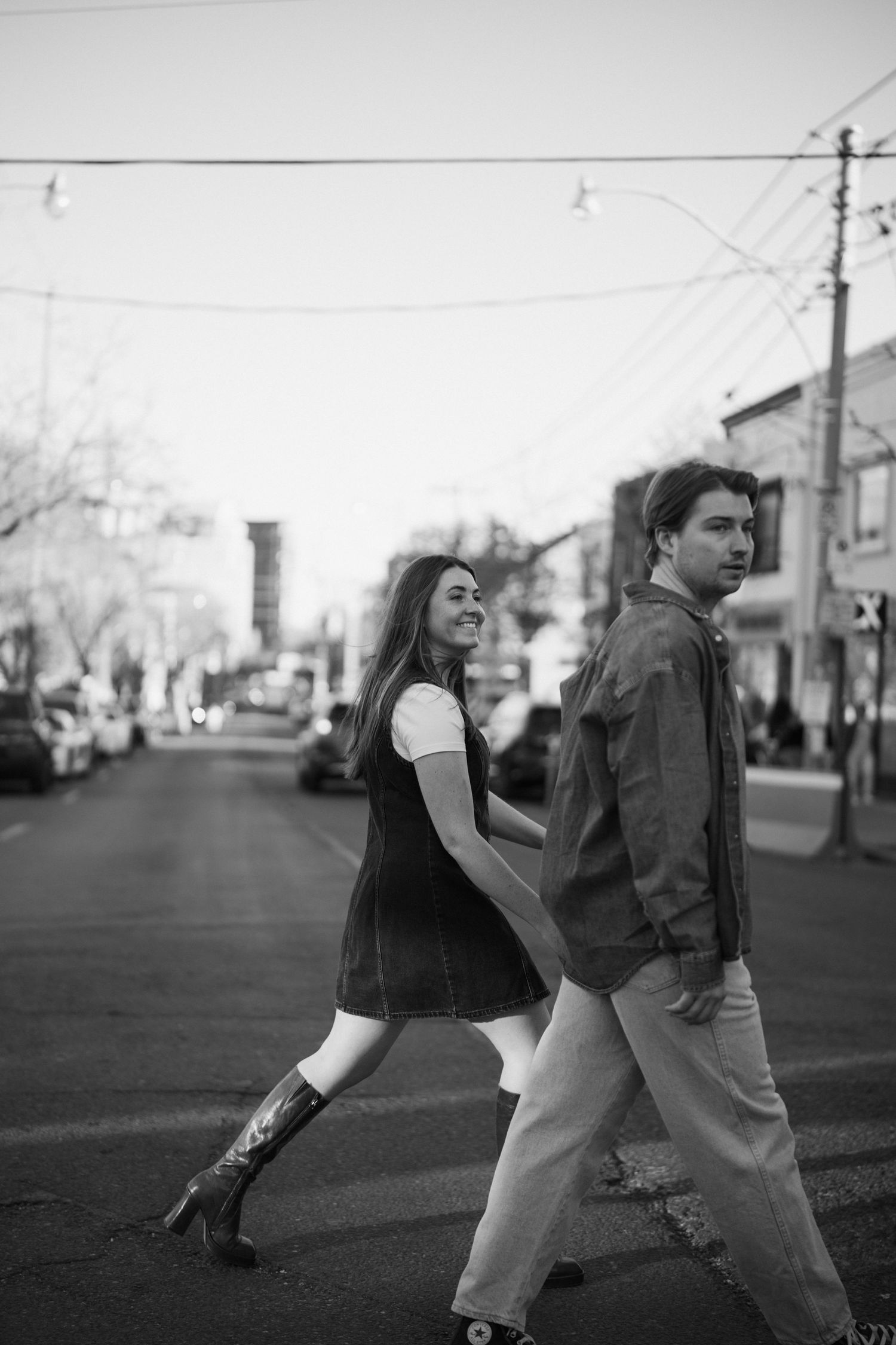 Black and white candid photo of a couple walking down a city street with utility poles and parked cars visible.