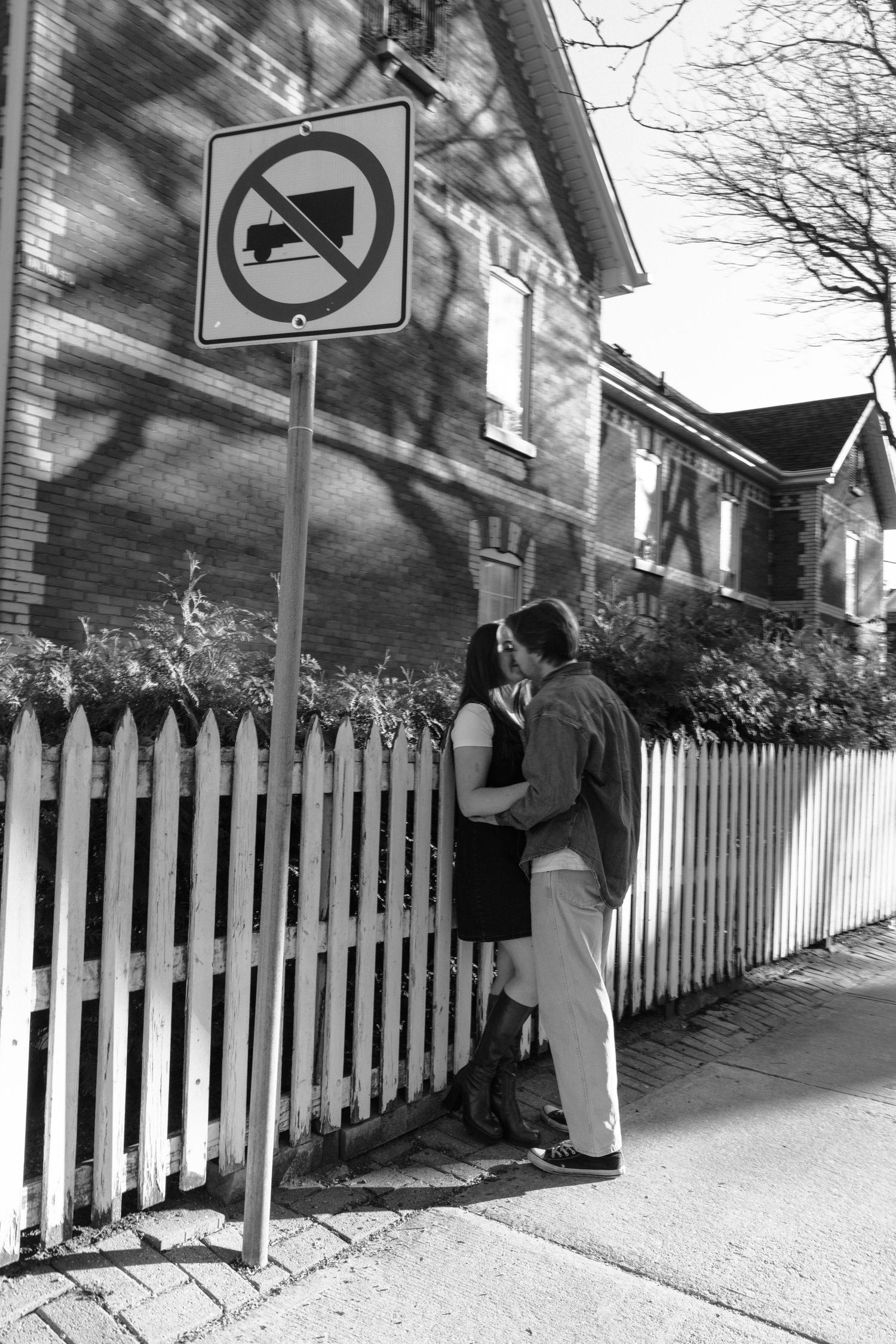 A couple shares a romantic moment by a white picket fence beneath a no parking sign on a residential street.