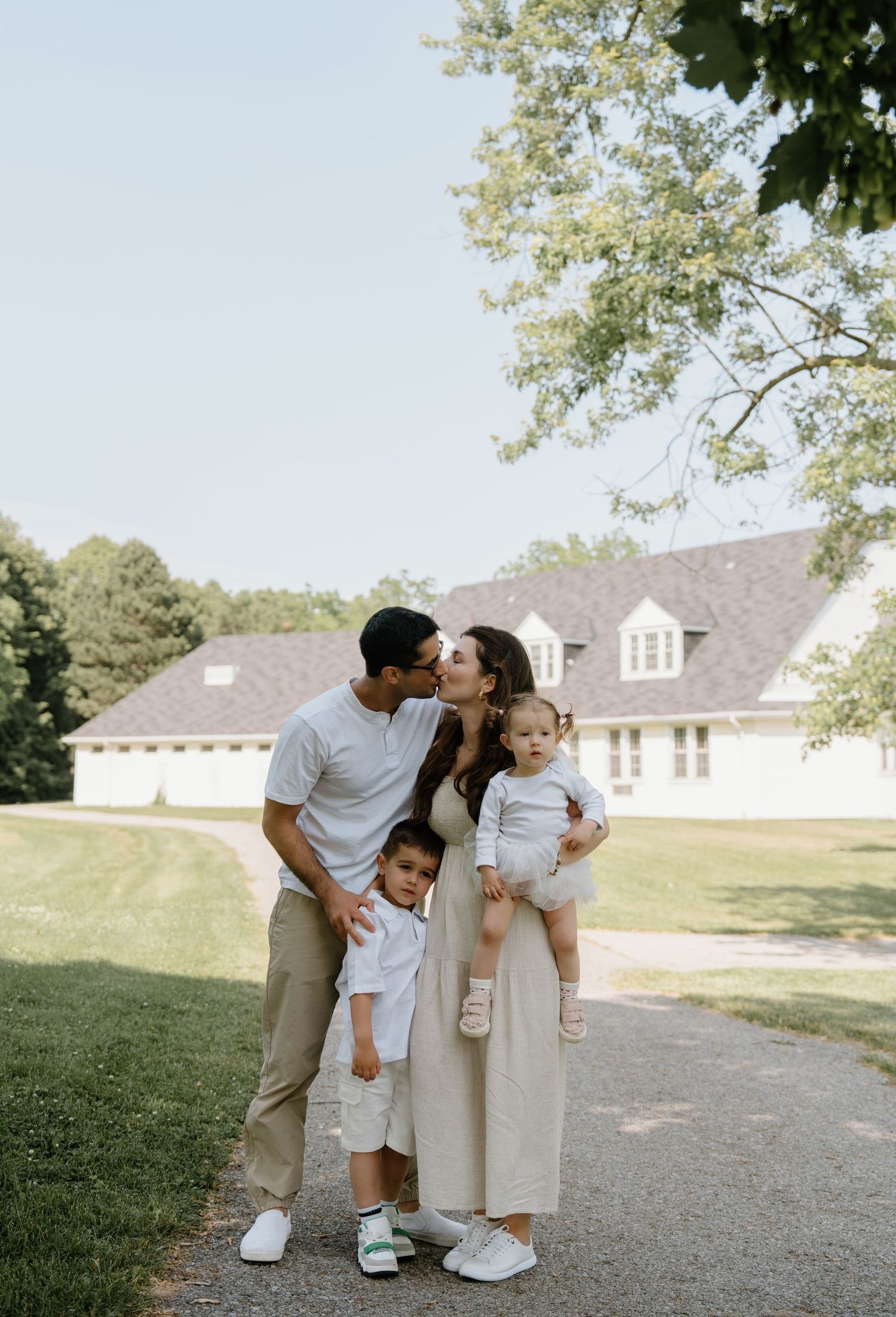 A family shares a tender moment together on a sunny day outside a white house with green lawn and trees.
