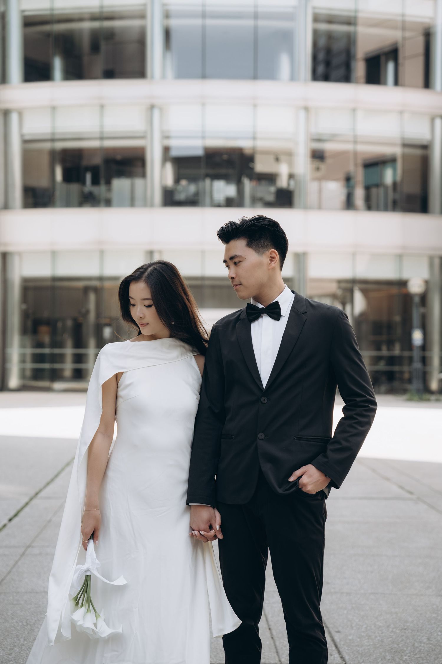 Elegant couple in formal wedding attire stands together outside modern building with white stone facade.