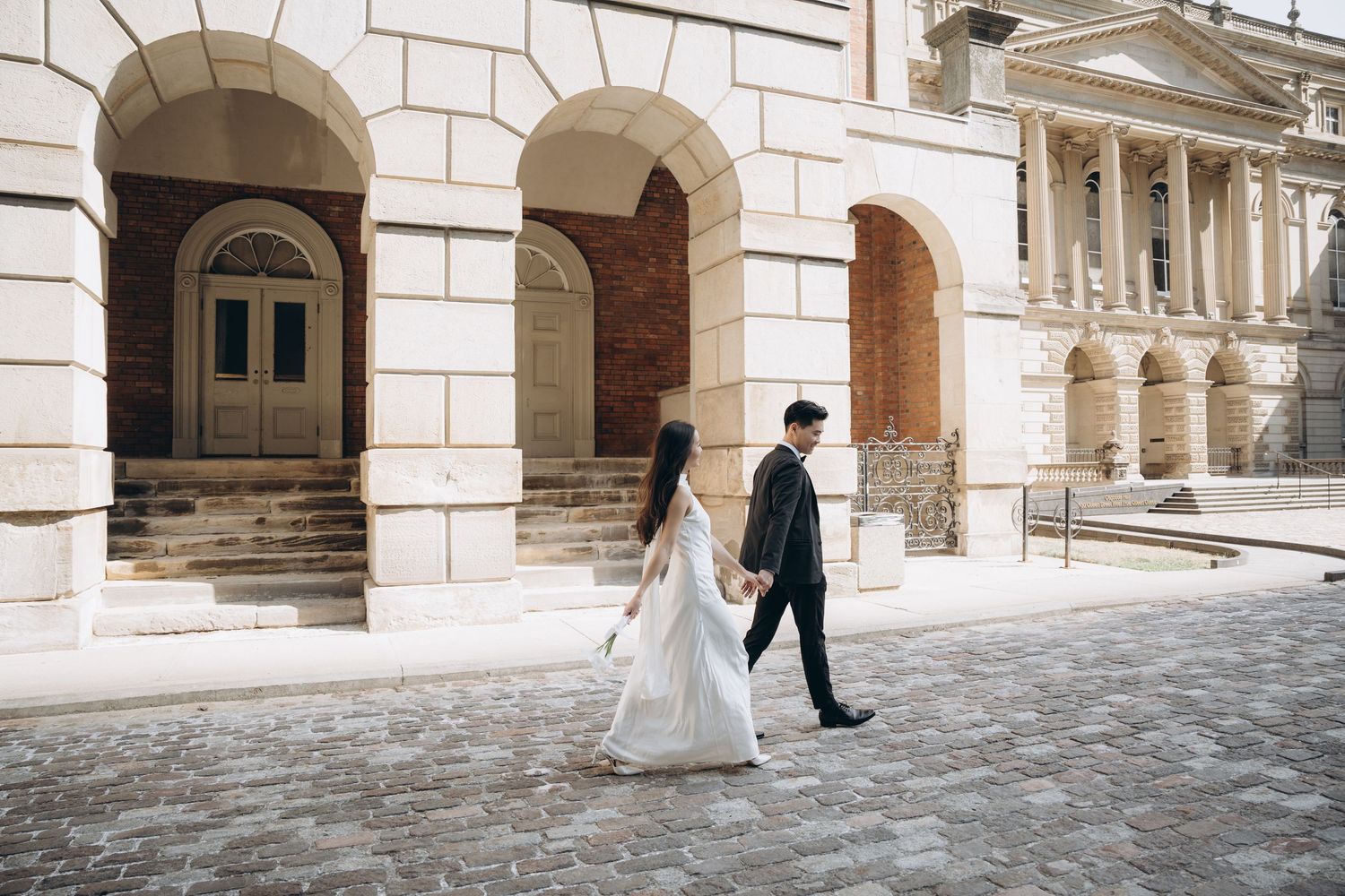 A couple in wedding attire walk along a snowy courtyard with elegant stone archways and classical architecture.