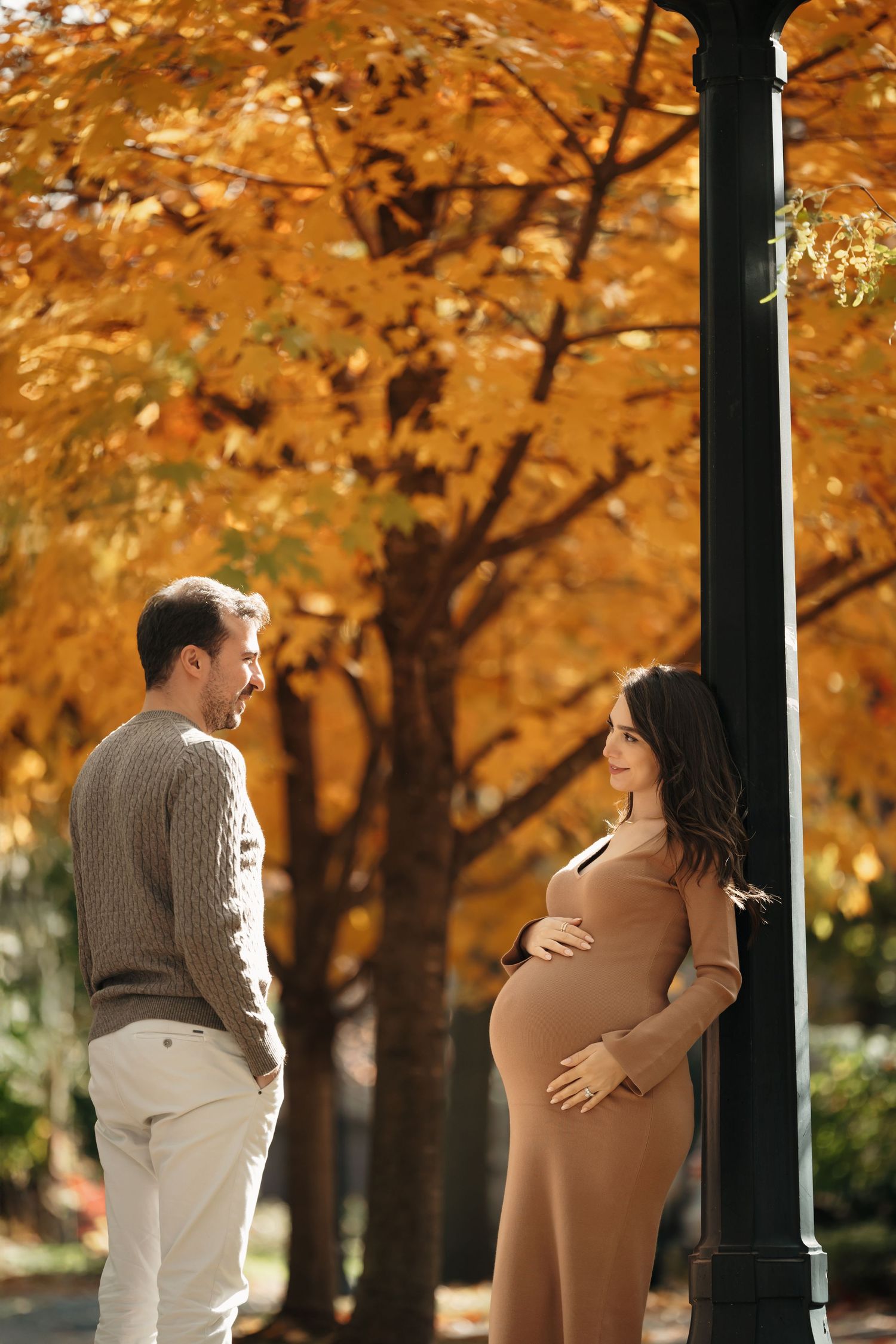 A couple in sweaters shares a tender moment outdoors during autumn with golden trees in the background.