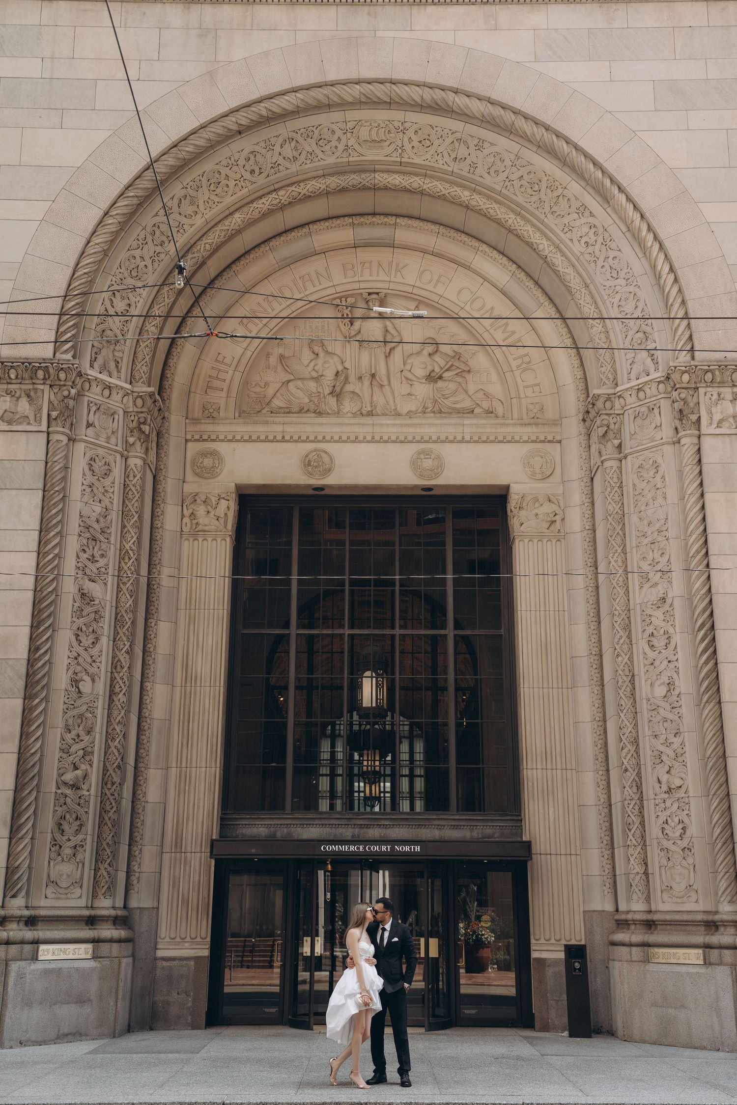 An elegant couple poses beneath ornate stone arches of a historic building's entrance.