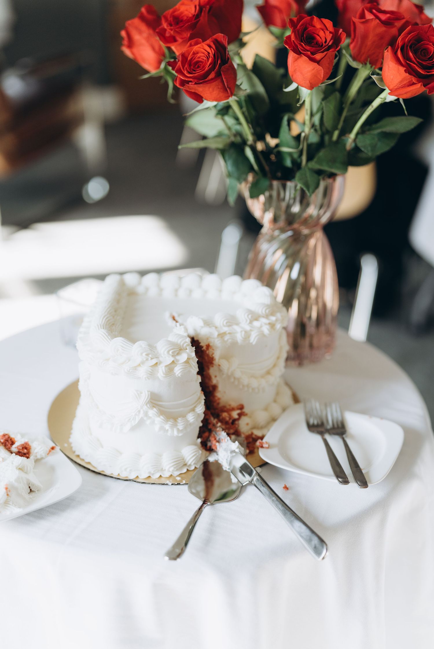 A close-up series of white wedding cake slices with red roses in crystal vases on a white tablecloth.