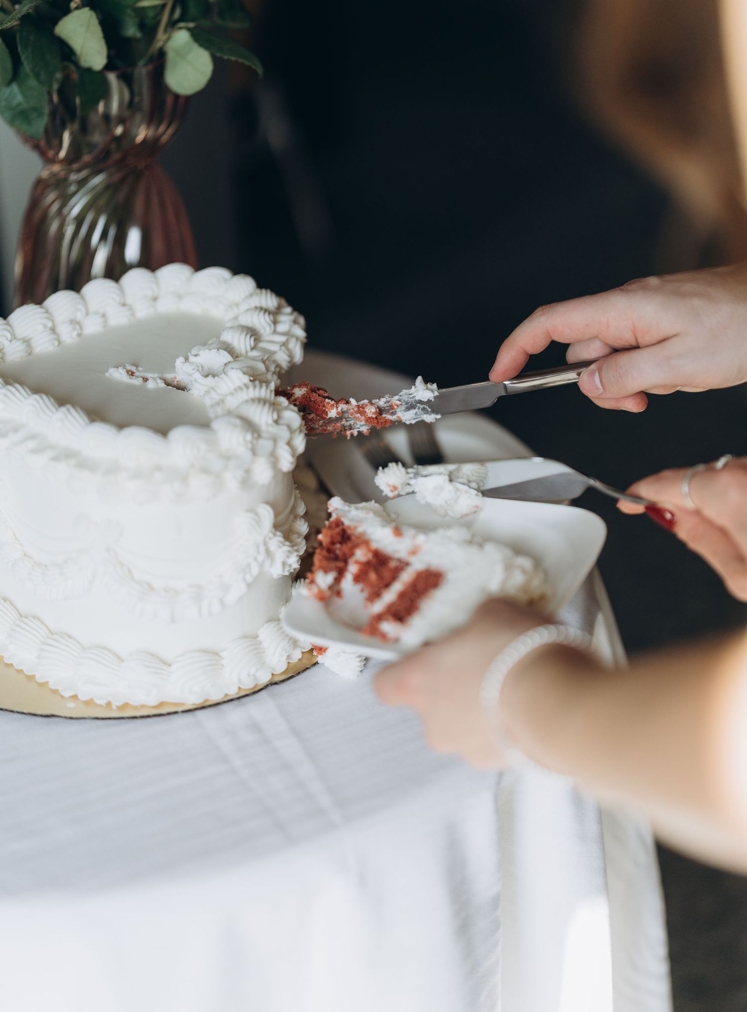 Two hands cut into a white layered wedding cake with decorative frosting using silver serving utensils.