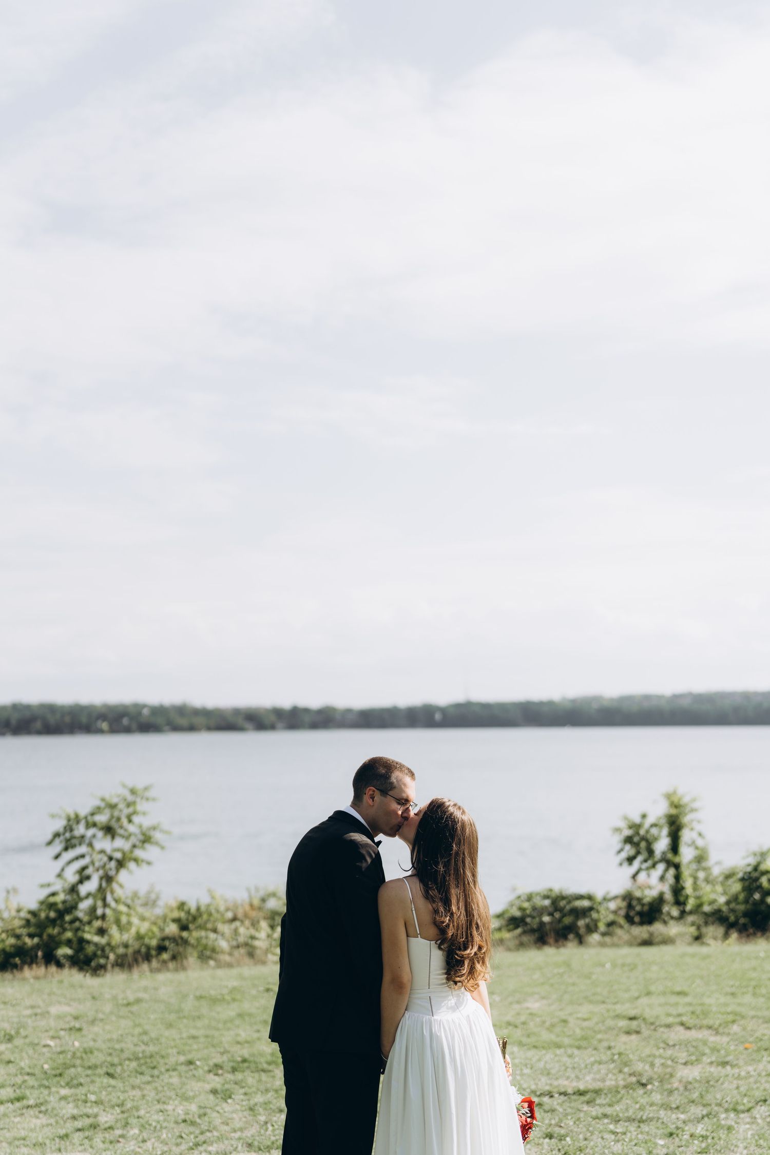 A romantic wedding couple stands together against a scenic lakeside backdrop during their outdoor ceremony.