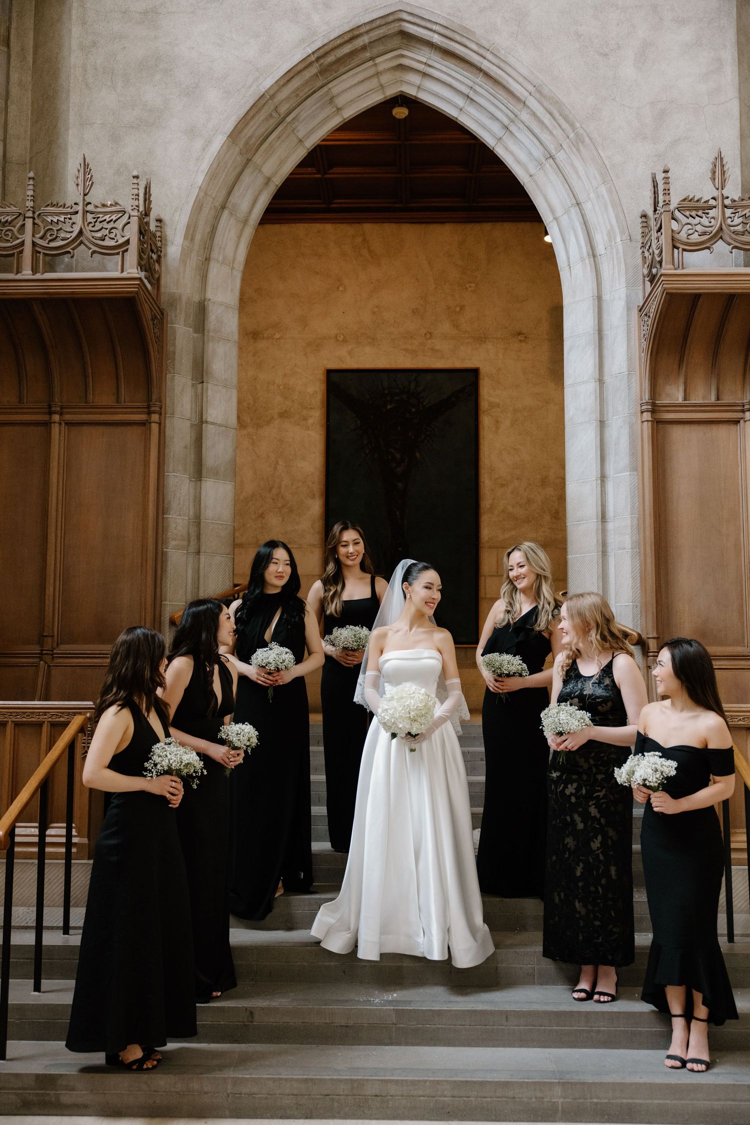 Wedding party in formal black dresses stands around bride in white gown on church steps under gothic stone archway.