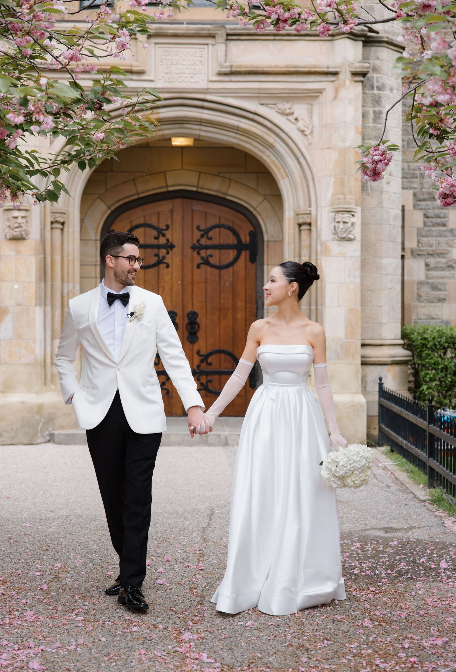 A newlywed couple in formal wedding attire holds hands while walking near an elegant stone archway with blooming trees.