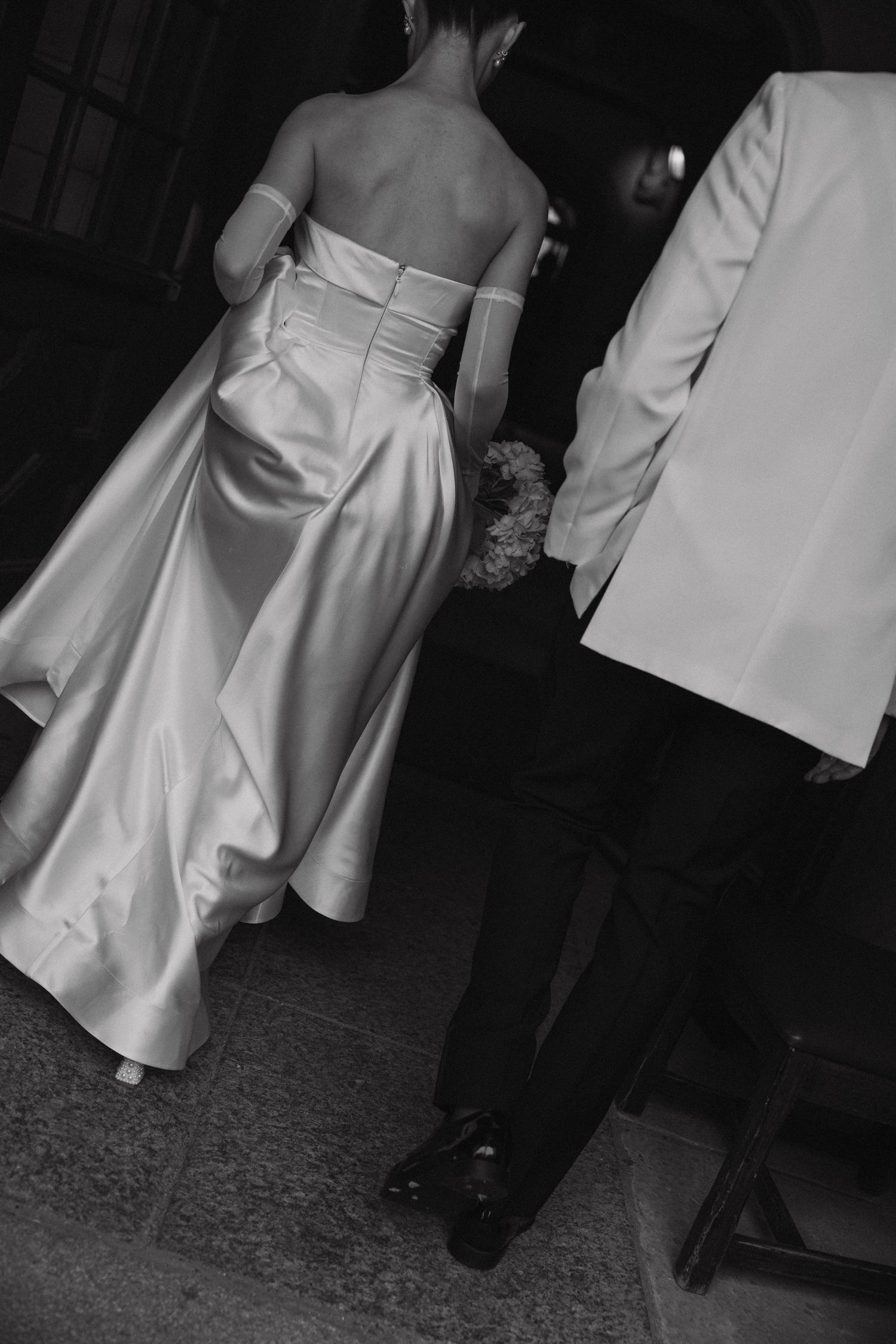 Back view of a bride in an elegant white satin wedding dress standing next to a person in a white suit jacket.