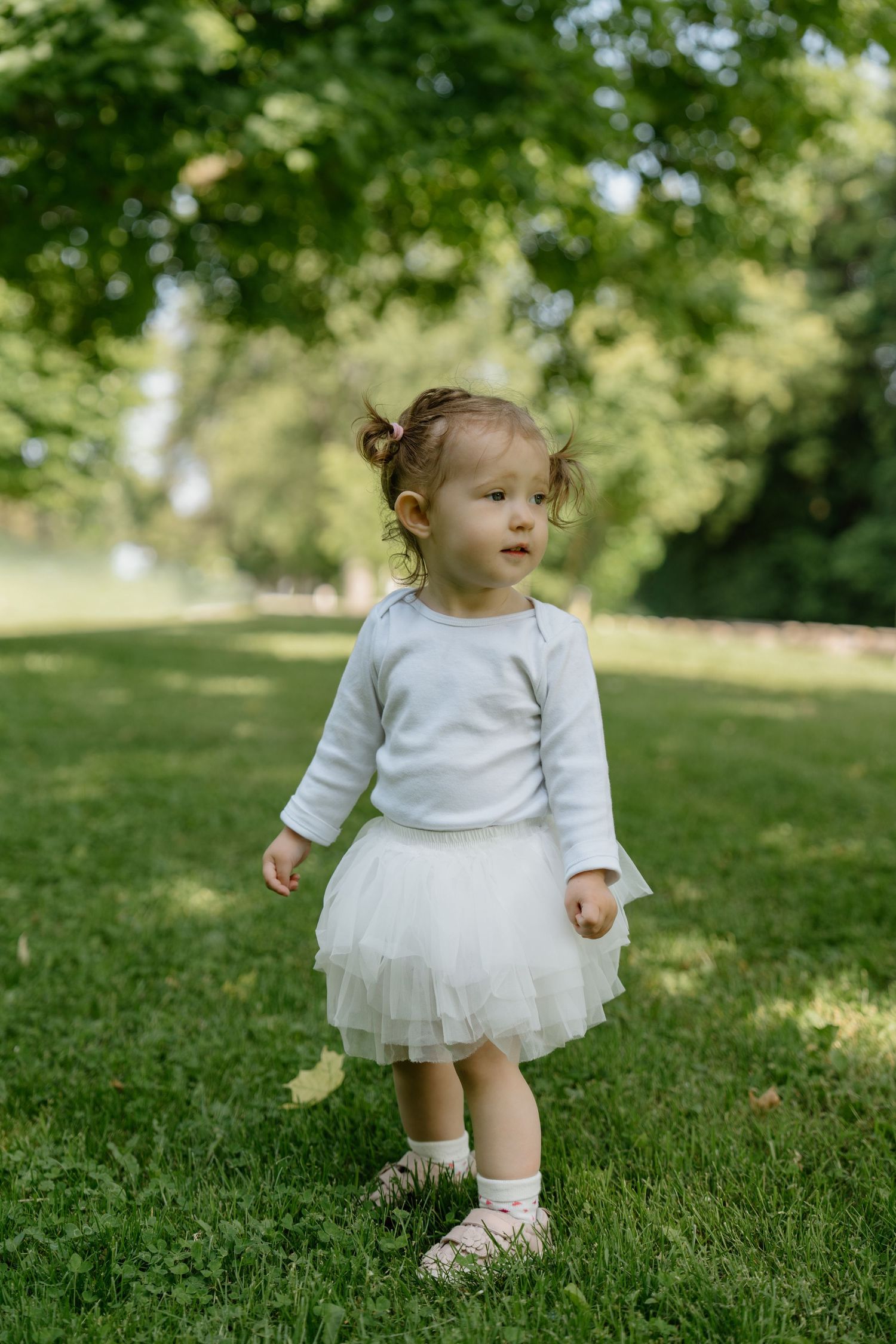 Small child wearing a white sweater and tulle skirt stands on grass in a sunny park.