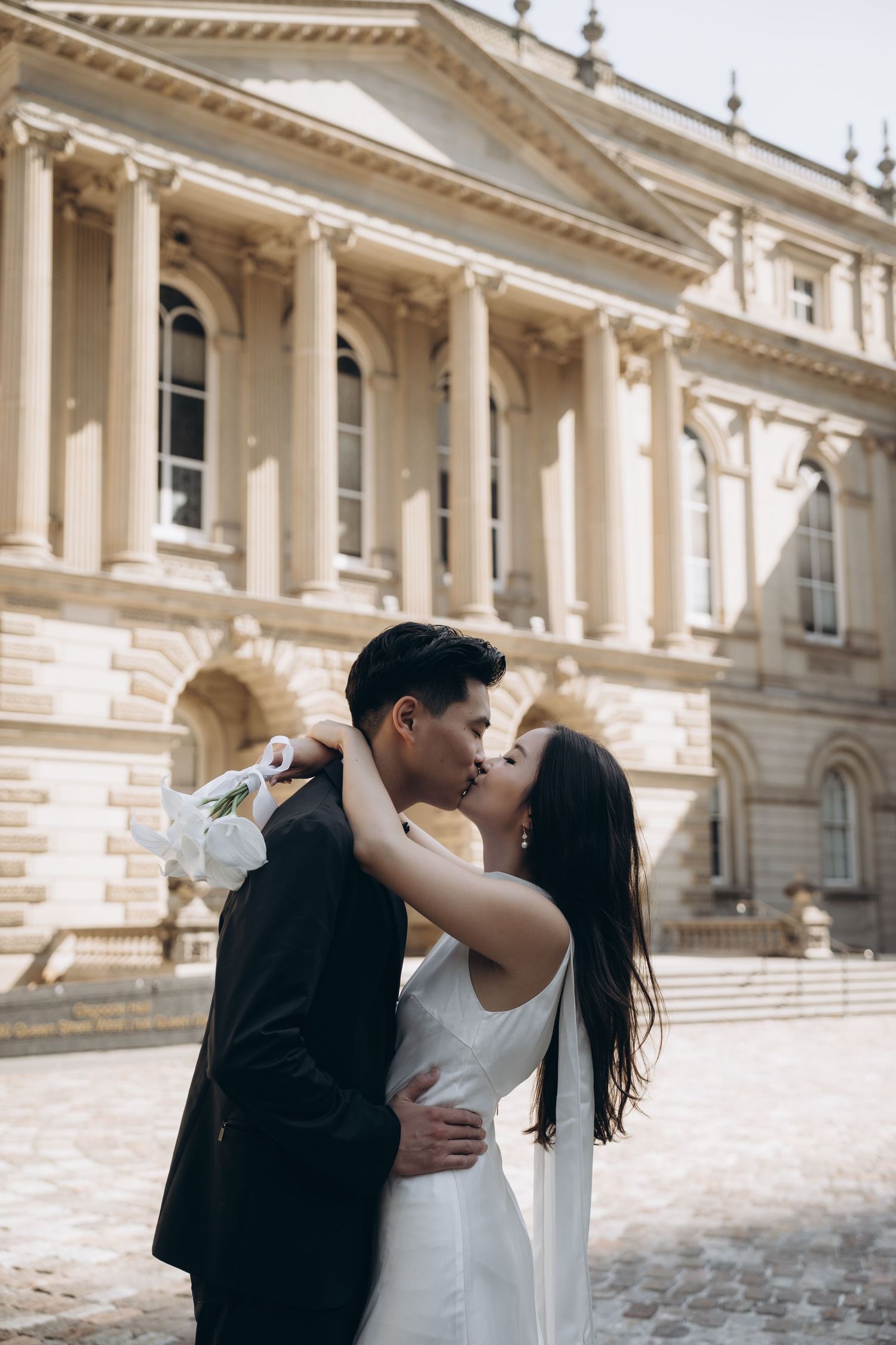 Romantic couple sharing intimate moments and kisses in front of ornate classical stone architecture.