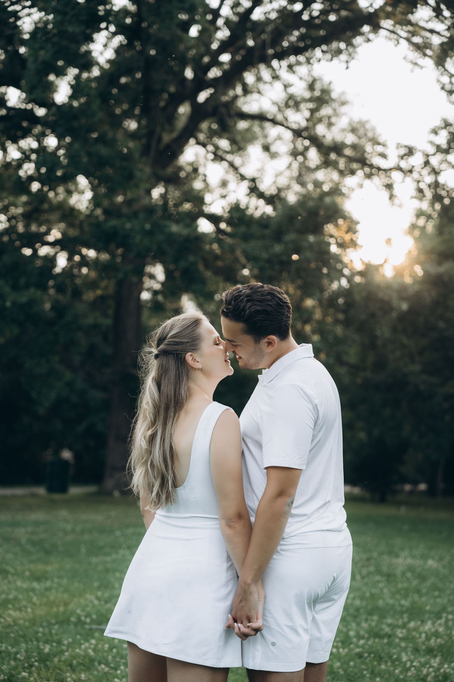 A couple dressed in matching white outfits share an intimate moment in a sunlit park setting.