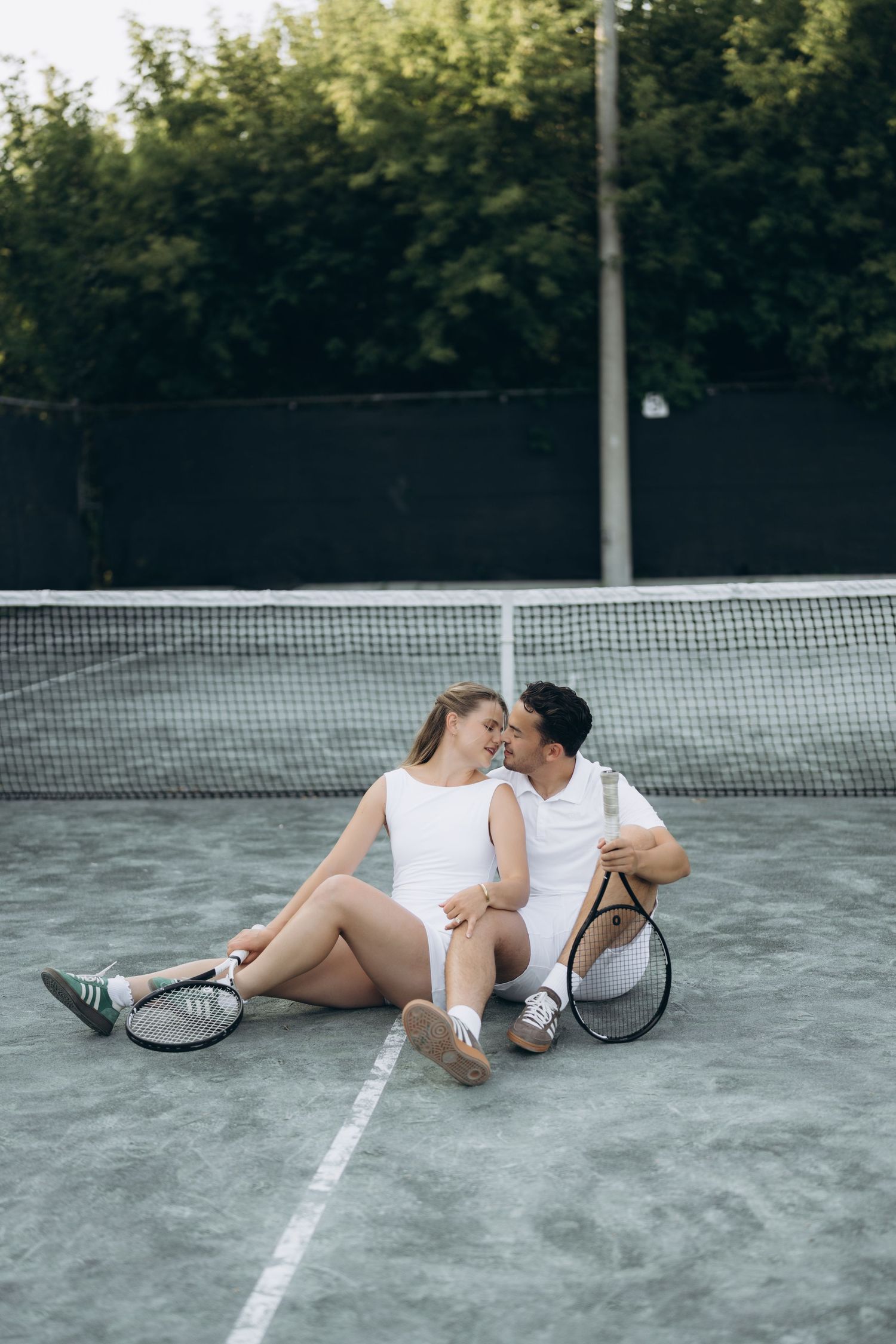 A couple in tennis whites sits together on a court next to the net while sharing an intimate moment.